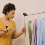 Mulher com bloco de notas e caneta na mão, olhando para um pequeno cabideiro com algumas peças de roupa, simbolizando o planejamento de um armário cápsula para economizar dinheiro e vestir-se melhor.