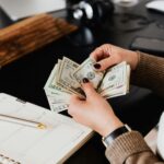 Mão de uma pessoa contando dinheiro sobre uma mesa, com um bloco de anotações, caneta e celular ao fundo, simbolizando planejamento financeiro e gestão de recursos.