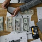 Mãos organizando notas de dinheiro ao lado de um smartphone com notificação de depósito e gráfico financeiro em ascensão desfocado ao fundo, simbolizando a gestão da restituição do Imposto de Renda 2025 para riqueza.