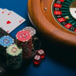 Roleta, fichas de cassino, baralho de cartas e notebook sobre uma mesa, representando o ambiente de jogos de azar online e as apostas esportivas.
